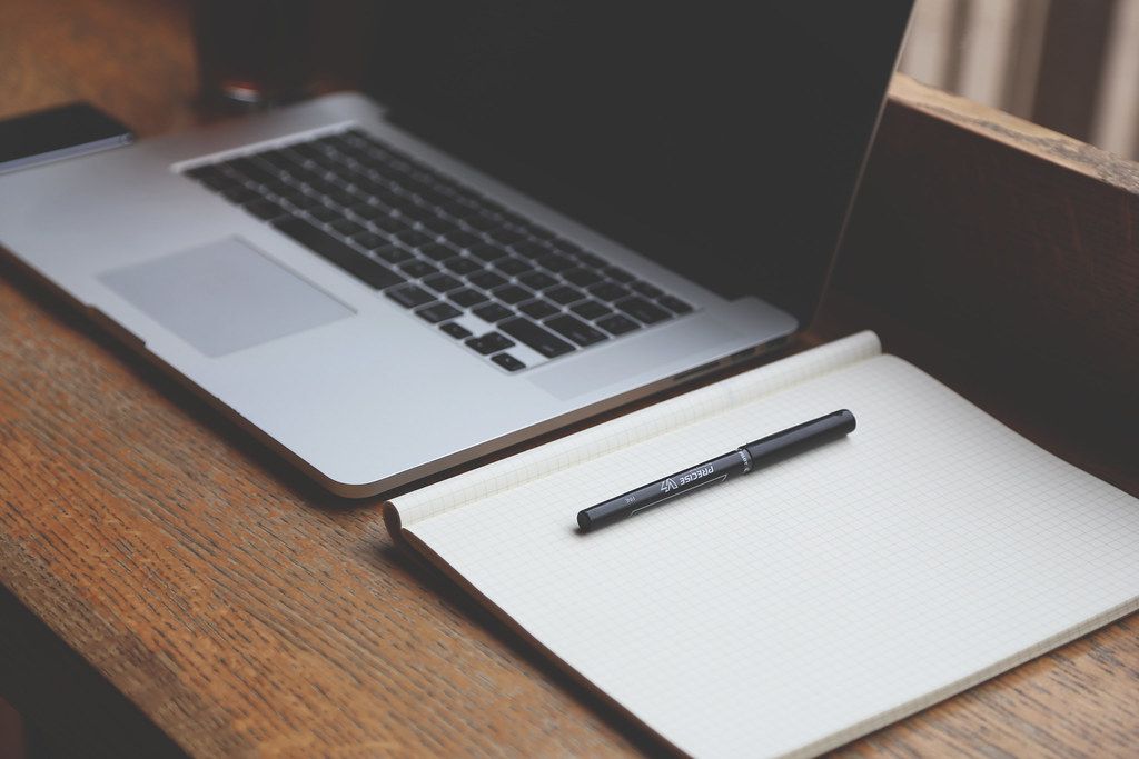 An open laptop beside a notebook and pencil on a wooden table.